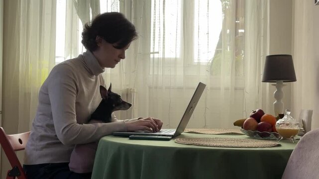 
Almaty, Kazakhstan - 02.02.2025 : A woman is sitting at her laptop, with her dog, in the kitchen. Work from home.