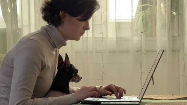 
Almaty, Kazakhstan - 02.02.2025 : A woman is sitting at her laptop, with her dog, in the kitchen. Work from home.