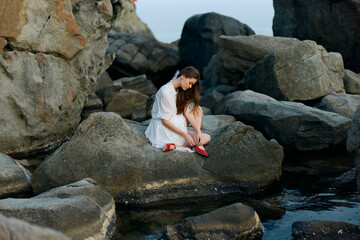 Woman in white dress sitting on ocean rocks with red shoes, gazing pensively at the horizon