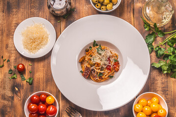 Spaghetti or Puttanesca pasta according to a traditional Italian recipe with anchovies, olives, capers on a white plate on a wooden background with ingredients on the table. Top view.