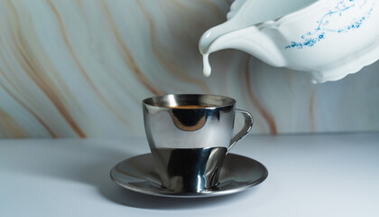 Silver cup filled with coffee as cream is poured from a porcelain pitcher on a marble surface