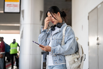 A worried Asian female traveler waits for her taxi at the airport pickup point, feeling headache.
