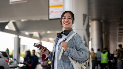 An attractive, smiling Asian female stands at an airport pickup point, waiting for a taxi.