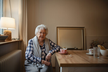 Lovely elderly woman sitting at the kitchen table and drinking a tea.