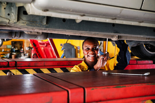 A smiling African female mechanic works under a lifted car in an auto repair shop. The scene highlights car maintenance, vehicle repair, and diversity in skilled trade professions.