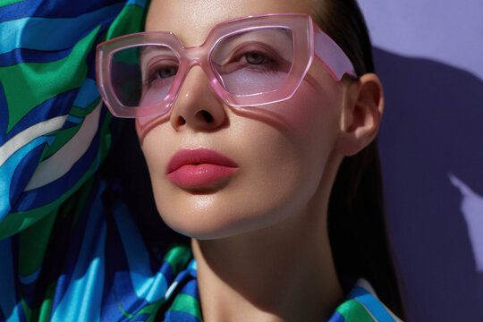 Fashion portrait of a woman wearing oversized pink sunglasses and a vibrant patterned blouse, with glossy lips and bold shadows..