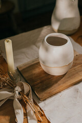 A candle and a vase are on a wooden table. The candle is lit and the vase is white. The table is covered with a white cloth