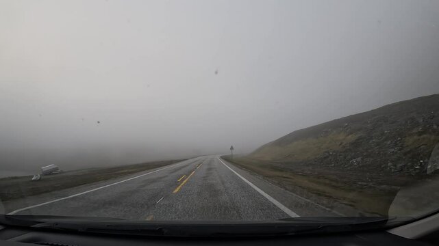 Driving on the E69 road through the tundra in northern Norway. Misty hills covered with grass and scattered stones surround the route, creating a remote and atmospheric landscape.