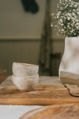 Two white ceramic bowls sit on a wooden cutting board. The bowls are empty and the cutting board is placed on a table