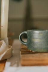 A green mug sits on a wooden board next to a candle. The candle is lit, casting a warm glow on the mug and the surrounding area. Concept of relaxation and comfort, as the candlelight
