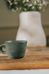A green coffee cup sits on a wooden table next to a white vase. The vase is filled with white flowers, giving the scene a fresh and inviting atmosphere. The combination of the coffee cup