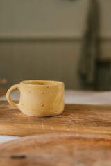 A yellow coffee cup sits on a wooden cutting board. The cup is small and has a handle. The cutting board is made of wood and is placed on a table. Concept of warmth and comfort