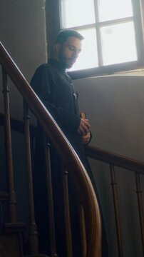 Vertical slow motion of gen Z pastor with bible book dressed in black soutane descending wooden stairs in church