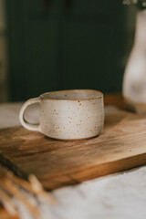 A white coffee cup sits on a wooden cutting board. The cup is plain and unadorned, with no design or pattern on it. The scene is simple and uncluttered, with the focus on the cup and the cutting board