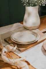 A white plate sits on a wooden board next to a vase of flowers. The scene is simple and elegant, with the white plate