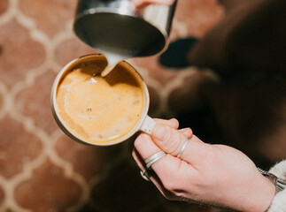 A person is pouring coffee into a cup. The coffee is brown and has a creamy texture. The person is holding the cup with both hands, and the coffee is being poured from a container