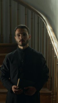 Vertical tilt up portrait of bearded gen Z pastor with holy book wearing black soutane confidently looking at camera with wooden staircase in background