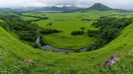 Fototapeta premium Winding river flows through a lush green valley surrounded by rolling hills and distant mountains under a cloudy sky.
