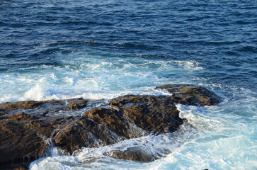Waves crashing on dark rocks along the Baiona coastline in Galicia, blending blue ocean hues with white foam, capturing the raw energy of the Atlantic

