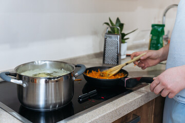 A person cooking on an induction stove, stirring grated carrots in a frying pan while a pot boils...
