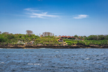 Picture a serene and tranquil scene of a lovely waterfront, dotted with lush greenery and charming homes, all set beneath a clear, bright blue sky filled with sunshine, Nusa Lembongan, Bali, Indonesia