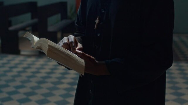 Midsection shot of unrecognisable priest dressed in dark clerical coat tracing lines with finger while reading bible book in catholic cathedral