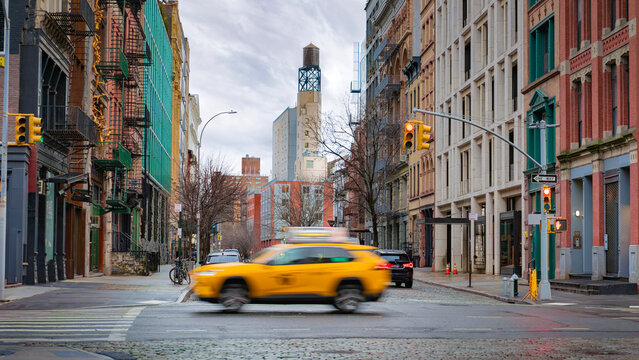 Busy New York City street with colorful historic buildings, a moving taxi, and a mix of urban details like cobblestones, fire escapes, and a water tower in the background