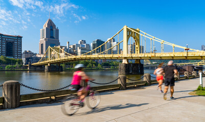 Lively scene in Pittsburgh, Pennsylvania, with downtown skyline in the background, iconic yellow bridge over Allegheny River, and two joggers and a cyclist motion blurred in the foreground