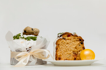 A festive Easter cake with white icing, decorated with green leaves, next to two vibrant yellow eggs and a wooden board, with a cut slice revealing a soft, fluffy texture with dried fruits inside.
