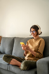 Happy smiling young student woman sitting on couch, listening music in headphones on mobile phone...