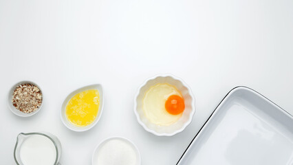 Flat lay composition for cooking preparation. Empty white baking dish with ingredients for baking, including egg, milk, butter, sugar, nuts