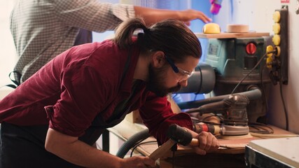 Naklejka premium Man carving intricate designs into wood using chisel and hammer in carpentry shop, enjoying diy hobby. Woodworking specialist in studio shaping wooden pieces with tools, making wood art, camera A