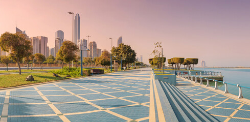 The Abu Dhabi Corniche street at sunrise offers a picturesque view of modern skyscrapers, the calm sea, and a beautifully designed promenade © EdNurg