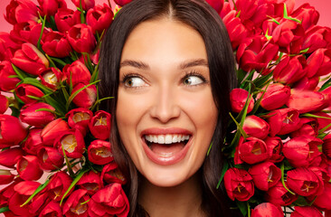 Excited woman posing with lots of red tulips flowers around her face, looking aside and smiling,...