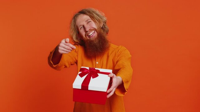 Positive smiling Caucasian man presenting birthday gift box stretches out hands offer wrapped present career bonus celebrating party promotion discount sale. Redhead guy isolated on orange background