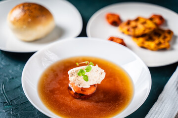 Soup or consomme, thick meat broth with carrots, chicken dumplings and microgreens, served in a white plate on a dark green background with sesame seed buns, napkin and spoon.