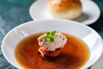 Soup or consomme, thick meat broth with carrots, chicken dumplings and microgreens, served in a white plate on a dark green background with sesame seed buns, napkin and spoon.
