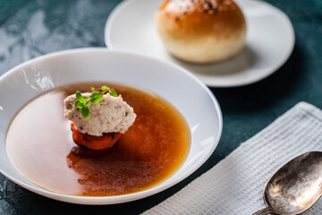 Soup or consomme, thick meat broth with carrots, chicken dumplings and microgreens, served in a white plate on a dark green background with sesame seed buns, napkin and spoon.