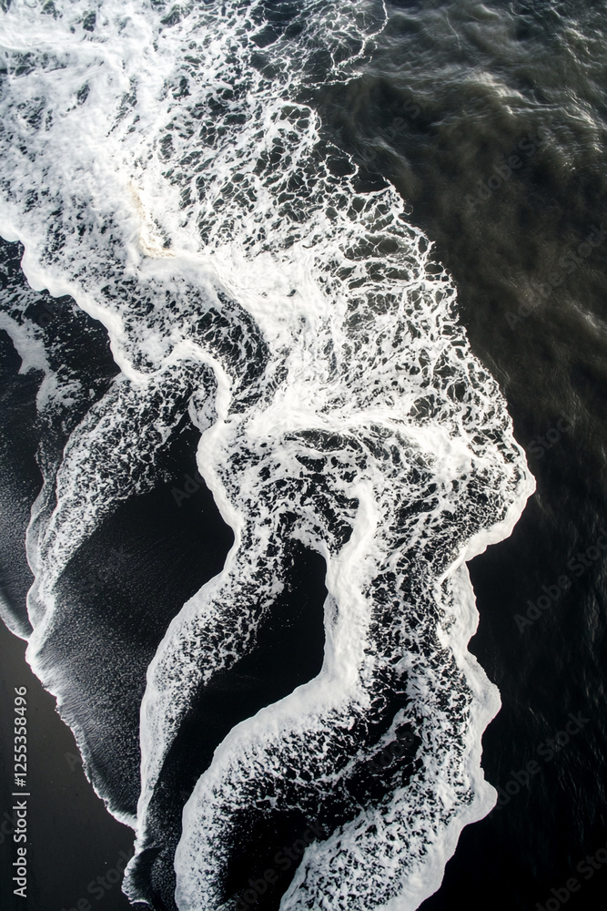 Poster aerial view of ocean waves crashing onto a black sandy beach, creating a dynamic contrast with white