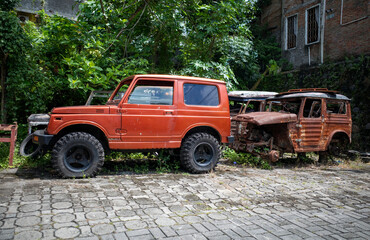 An old red car and a rusty older generation car without wheels