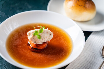 Soup or consomme, thick meat broth with carrots, chicken dumplings and microgreens, served in a white plate on a dark green background with sesame seed buns, napkin and spoon.
