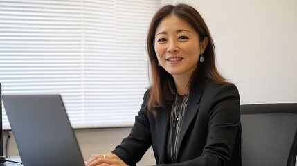 A young woman in business attire is smiling and using a laptop at a desk with a white background