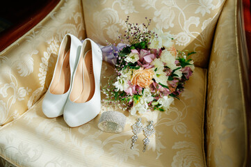 Elegant wedding setup featuring white bridal shoes, a pastel floral bouquet, crystal earrings, and...