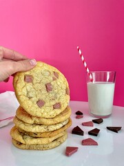 hand takes a cookie from a cookie tray near a glass of milk on a pink background