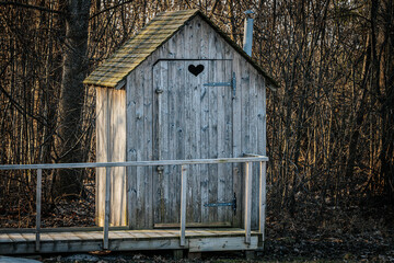 A weathered wooden outhouse with a heart-shaped cutout on the door, located in a forested area. A wooden walkway with railings leads to the entrance, surrounded by bare trees.