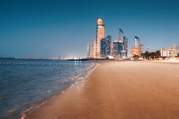 Naklejka premium Illuminated skyscrapers of Abu Dhabi reflecting on the wet sand of the beach at dusk, creating a vibrant, tranquil cityscape by the sea