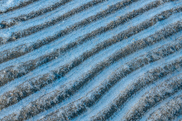 Close-up of sand ripples with a dusting of frost, creating a textured and wavy pattern. The contrast between dark sand and icy blue tones highlights natural formations.