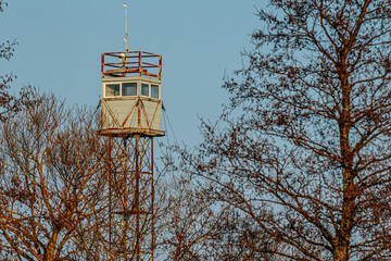 A rusty old watchtower with a weathered metal frame stands against a clear blue sky, with bare tree...