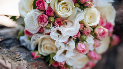 Close-up of a wedding bouquet featuring pink and white roses, with gold wedding rings placed on top, resting on a rustic surface.  
