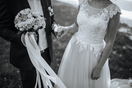 Black and white image of a bride in a lace wedding dress and groom holding hands, with a bouquet of roses tied with ribbons.  
